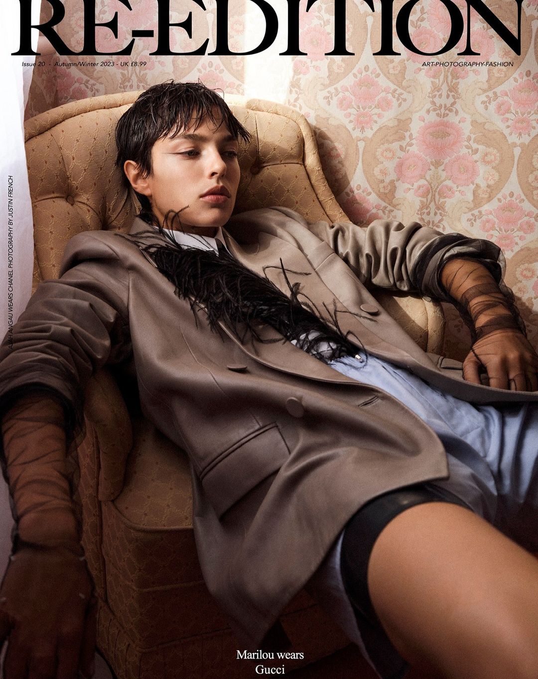 Model with wet-look hair seated in an armchair wearing an oversized blazer against vintage patterned wallpaper.