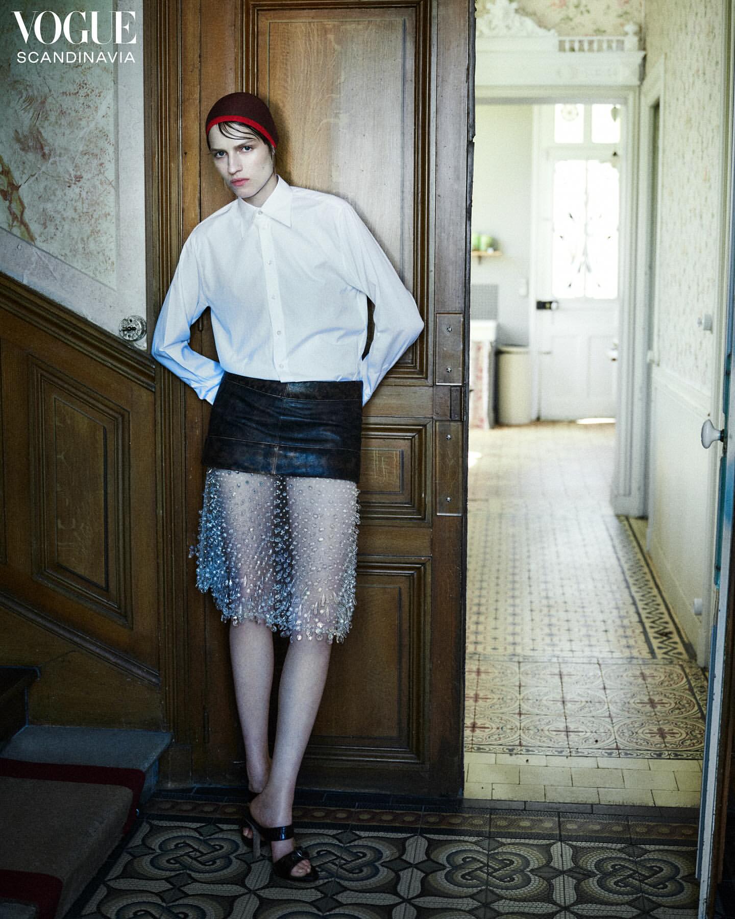 Fashion editorial shot of a young adult woman in white shirt, mini skirt, and red cap standing in front of wooden doorway in hallway.