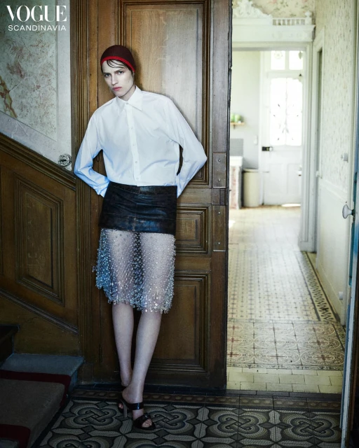Fashion editorial shot of a young adult woman in white shirt, mini skirt, and red cap standing in front of wooden doorway in hallway.