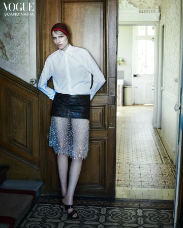 Fashion editorial shot of a young adult woman in white shirt, mini skirt, and red cap standing in front of wooden doorway in hallway.