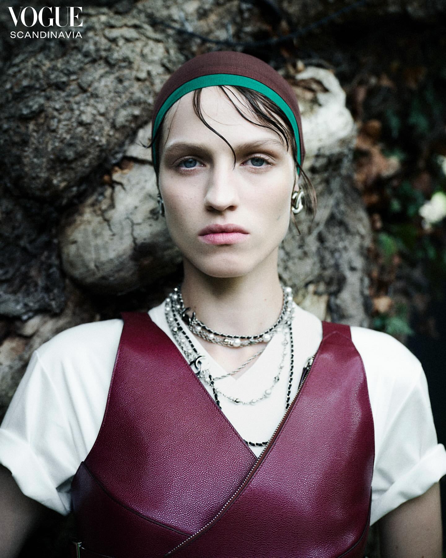 Close-up of a woman with dark blonde hair and a brown headscarf with a green band, wearing a red leather top and layered necklaces.