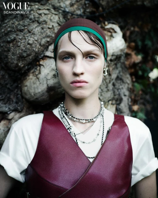 Close-up of a woman with dark blonde hair and a brown headscarf with a green band, wearing a red leather top and layered necklaces.