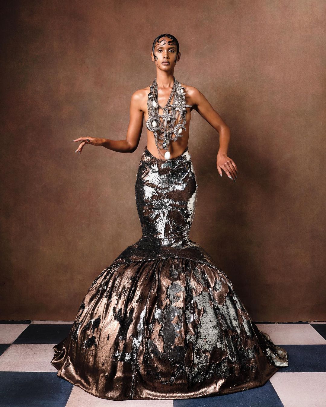 Adult female model posing in a textured, bronze-and-silver mermaid gown with an elaborate silver necklace and headpiece against a brown backdrop.
