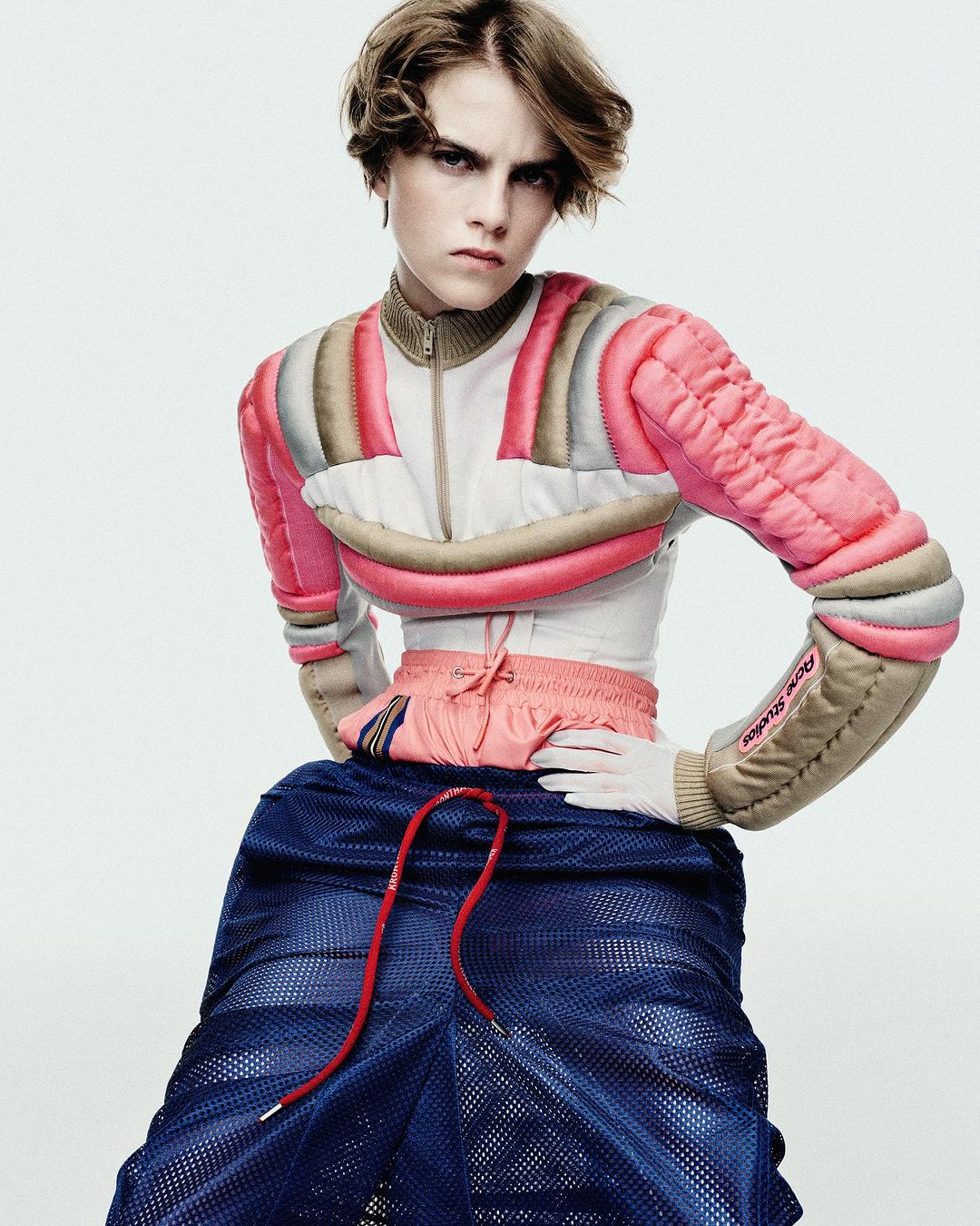 An androgynous person poses in a light studio wearing a pink and beige padded crop top and blue mesh skirt, with a stern facial expression.