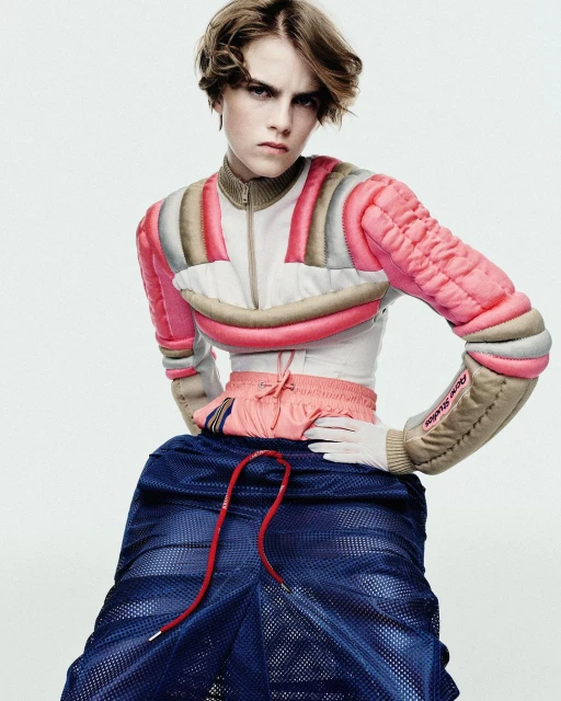 An androgynous person poses in a light studio wearing a pink and beige padded crop top and blue mesh skirt, with a stern facial expression.
