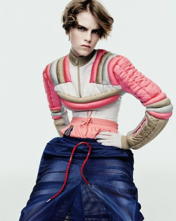 An androgynous person poses in a light studio wearing a pink and beige padded crop top and blue mesh skirt, with a stern facial expression.