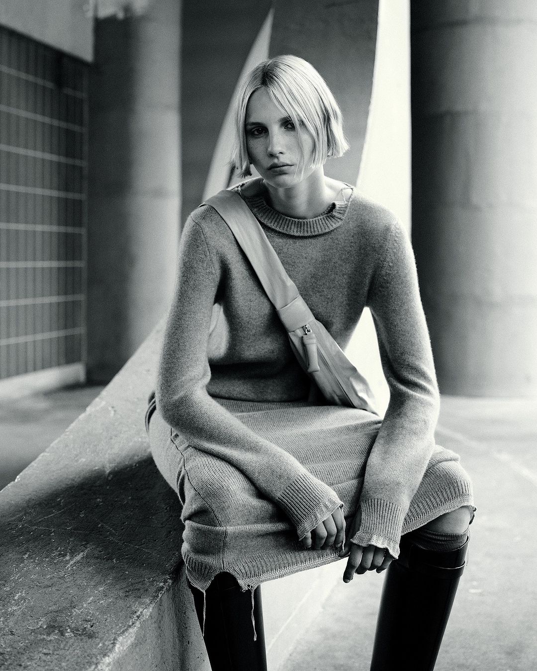 A woman in a gray sweater dress and black boots sits against a concrete background wearing a shoulder bag with a neutral expression.
