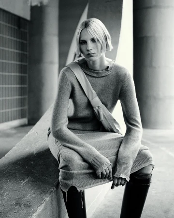 A woman in a gray sweater dress and black boots sits against a concrete background wearing a shoulder bag with a neutral expression.
