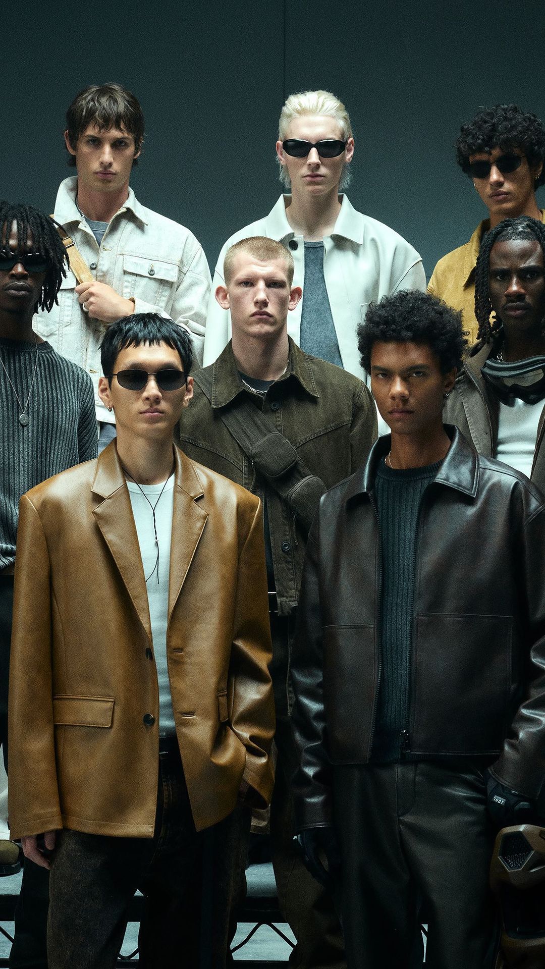 Group portrait of nine men in a studio setting, wearing fashionable clothing, posing with neutral expressions under studio lighting.
