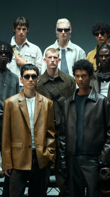 Group portrait of nine men in a studio setting, wearing fashionable clothing, posing with neutral expressions under studio lighting.