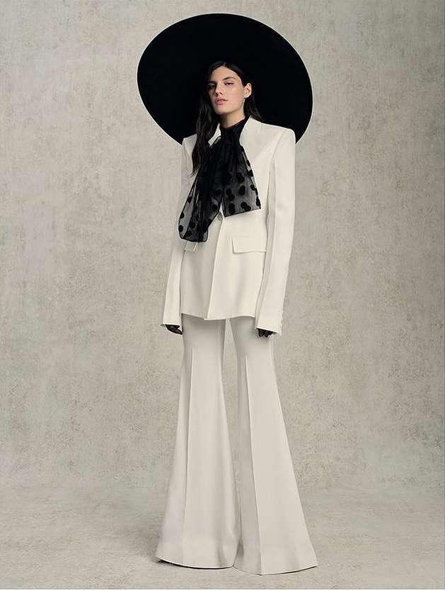 Elegant woman in a white pantsuit with a black polka dot scarf and large black hat posing against a neutral textured backdrop.