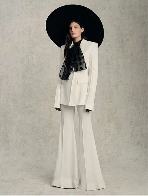 Elegant woman in a white pantsuit with a black polka dot scarf and large black hat posing against a neutral textured backdrop.