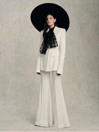 Elegant woman in a white pantsuit with a black polka dot scarf and large black hat posing against a neutral textured backdrop.