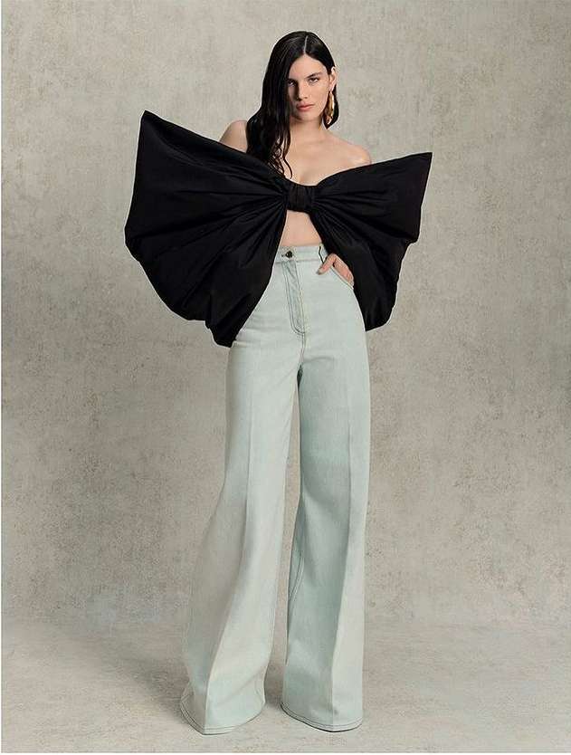 Fashion shot of a woman in wide-leg light blue pants and a large black bow top standing indoors against a neutral background.