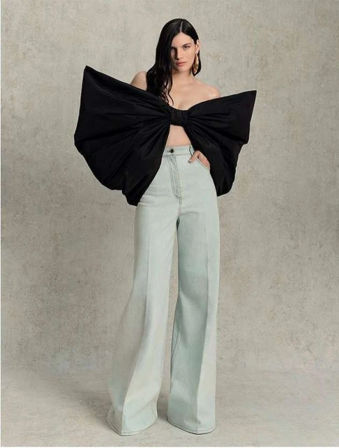 Fashion shot of a woman in wide-leg light blue pants and a large black bow top standing indoors against a neutral background. 