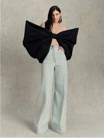 Fashion shot of a woman in wide-leg light blue pants and a large black bow top standing indoors against a neutral background. 