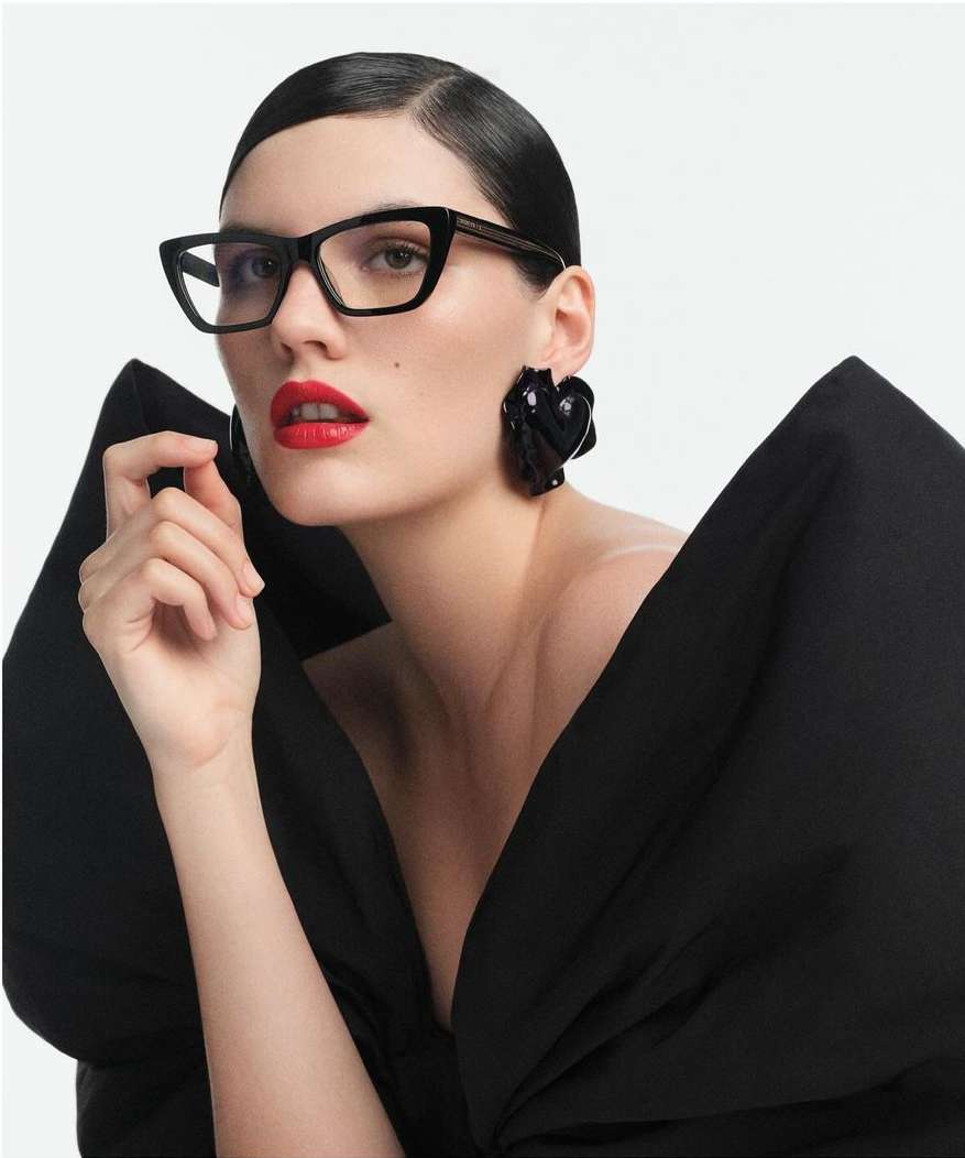 Woman wearing black cat-eye glasses and large sculptural earrings with bright red lipstick against a plain white background