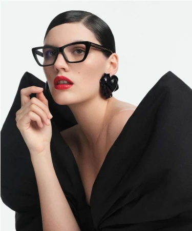 Woman wearing black cat-eye glasses and large sculptural earrings with bright red lipstick against a plain white background
