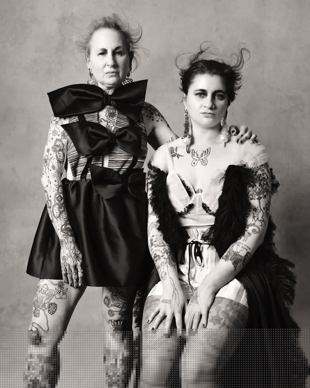 Two women pose in a photo studio, adorned with tattoos and elaborate clothing. One wears a dress with large black bows.