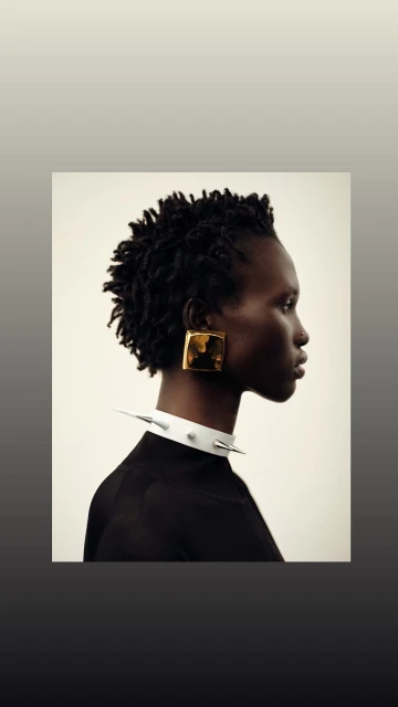 Profile view of Black woman with natural hair, gold square earrings, black top, and spiked white collar against a white backdrop.