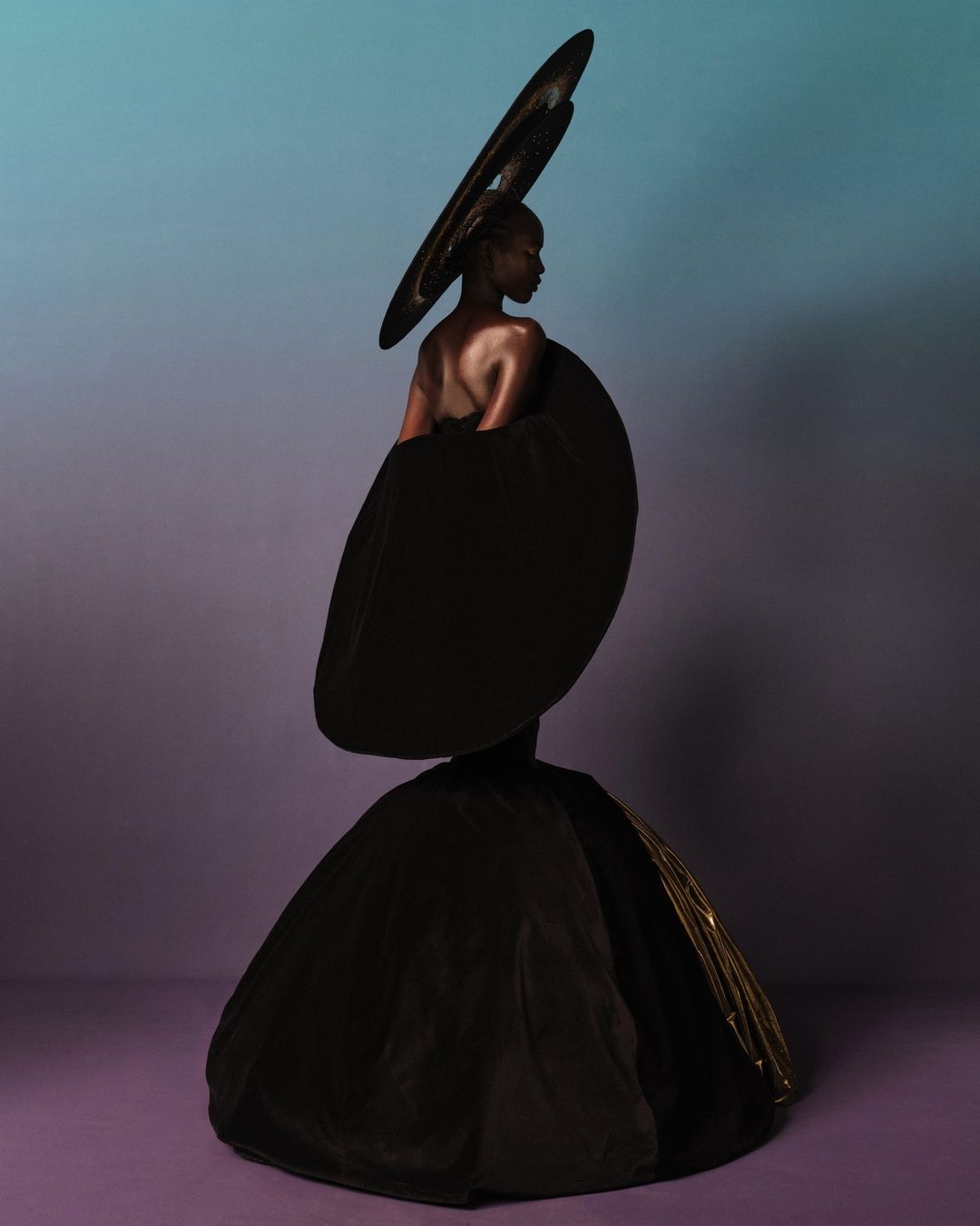 Model wearing dramatic sculptural black gown and oversized circular headpiece against a gradient studio background.