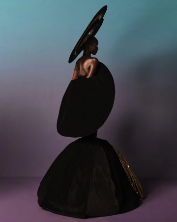 Model wearing dramatic sculptural black gown and oversized circular headpiece against a gradient studio background.