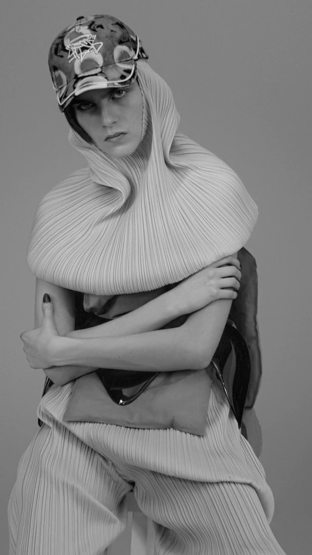 Monochrome fashion portrait of a female model wearing a textured pleated outfit with a baseball cap in a studio setting. 