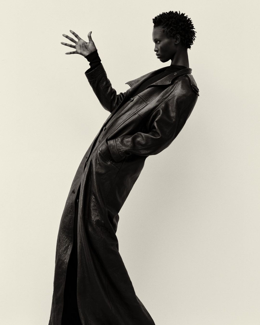 Black woman in long black leather coat posing dramatically with outstretched hand against a light backdrop