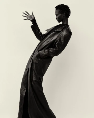 Black woman in long black leather coat posing dramatically with outstretched hand against a light backdrop