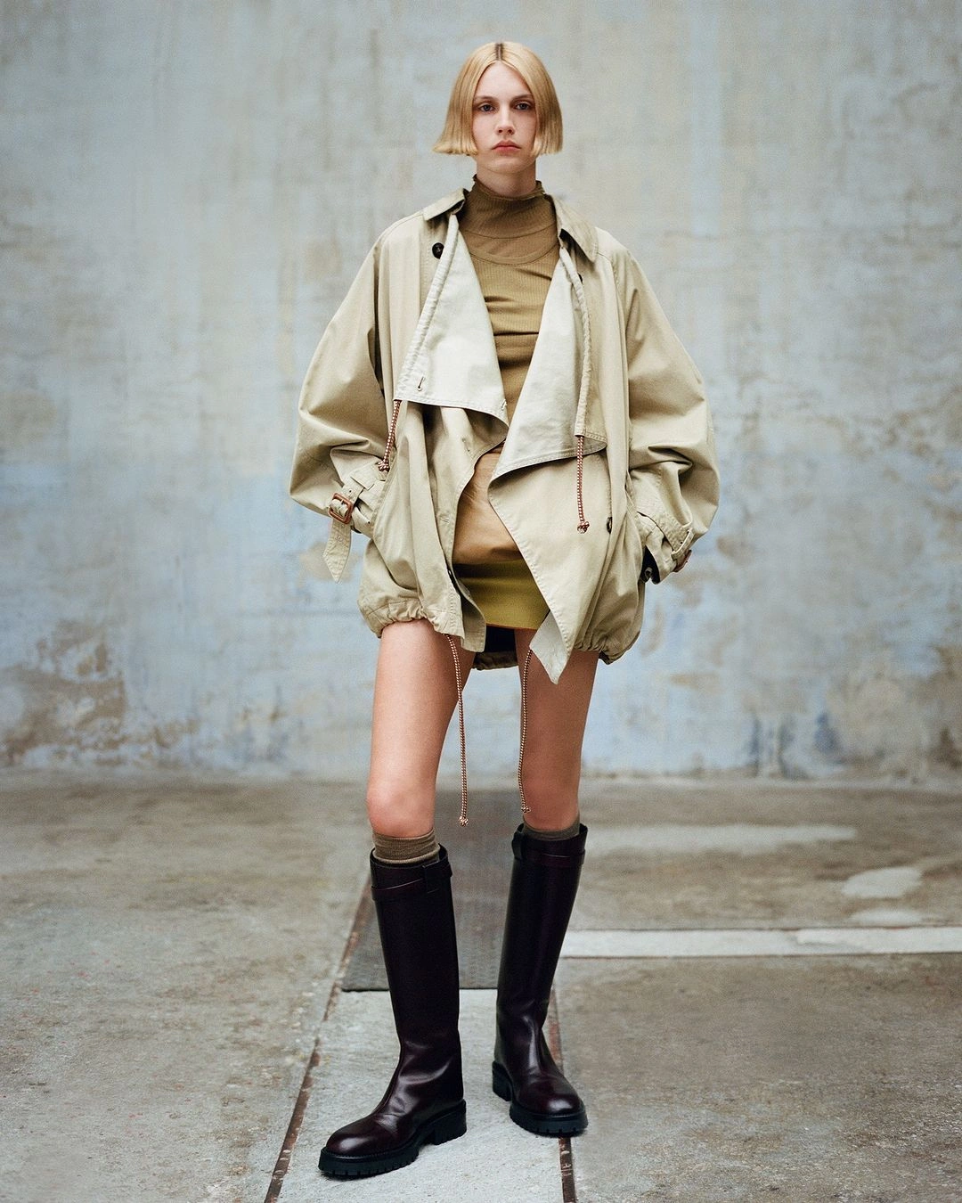 Model with short blond hair wearing an oversized beige jacket and dark knee-high boots against a weathered concrete wall.