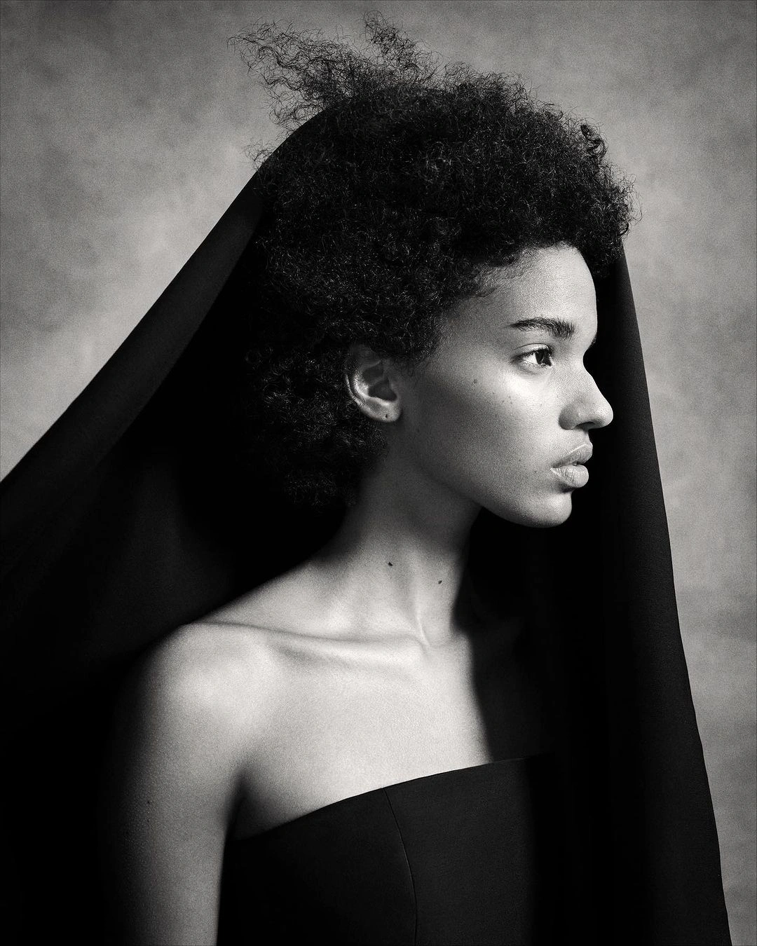 Monochrome portrait of a black woman in profile with a dark head covering and strapless black top against a mottled grey backdrop.