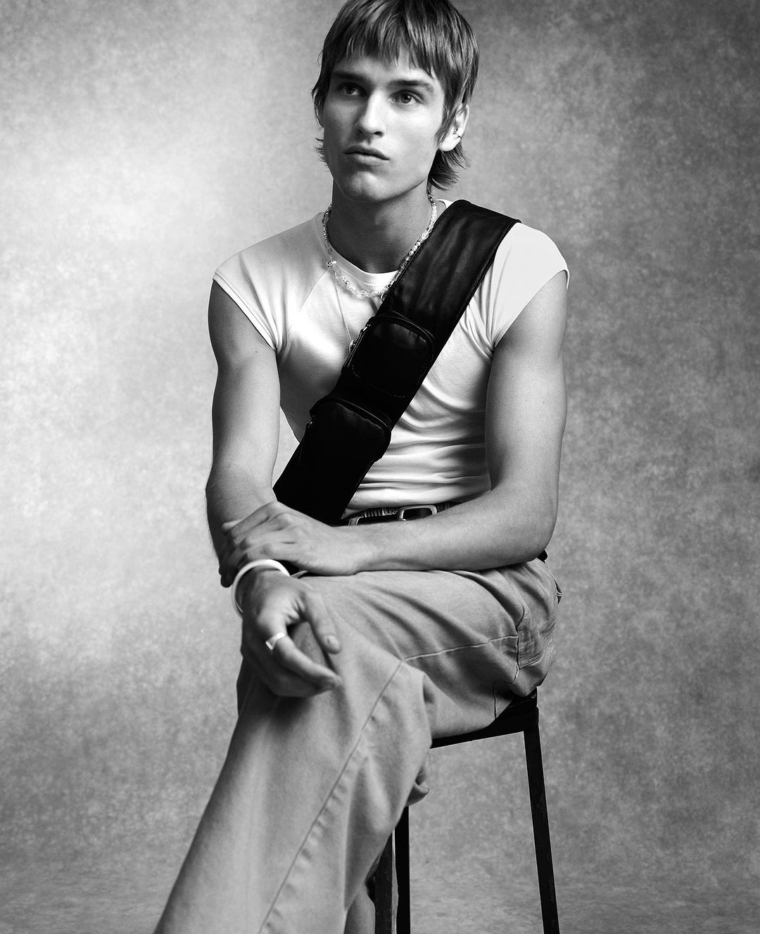 Young male model with layered hair wearing a white t-shirt and crossbody bag seated on a stool against a mottled grey background.