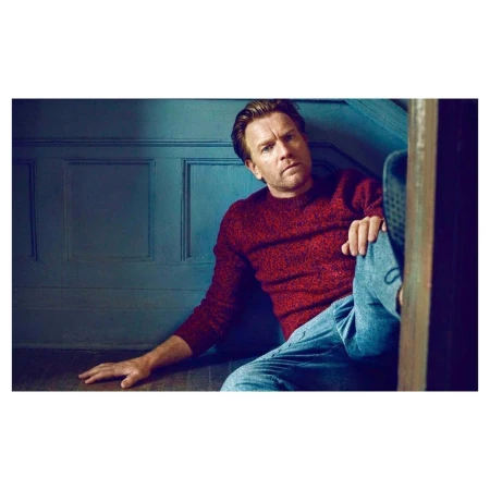 Actor Ewan McGregor wearing a red textured sweater and blue jeans sitting against a blue paneled wall.