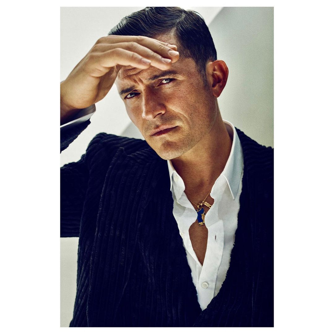 Orlando Bloom portrait in a corduroy suit with a white shirt looking pensively at the camera with hand on his forehead.