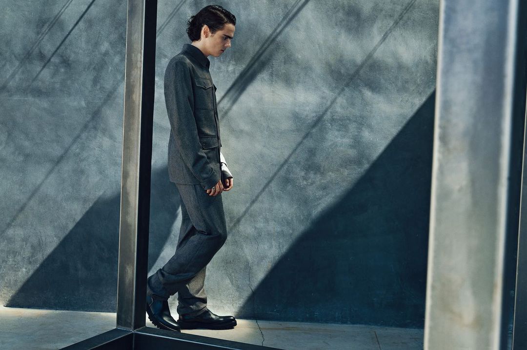 A young adult male is walking in a gray suit in front of a concrete wall with shadows. Fashion shot in studio lighting.
