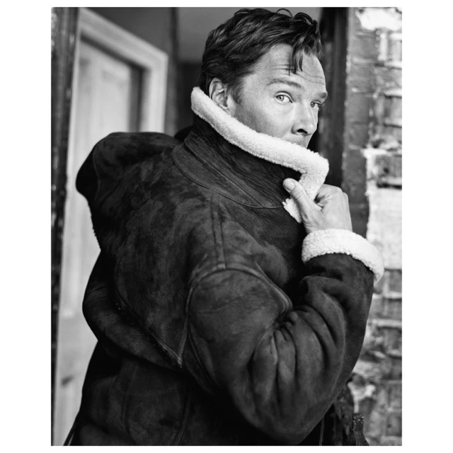Black and white portrait of a man wearing a coat with a fur collar, looking over his shoulder, standing beside a brick wall.