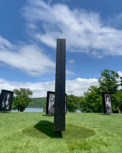 Outdoor art installation featuring black monolith and framed portraits on green grass under a bright blue sky with scattered clouds.