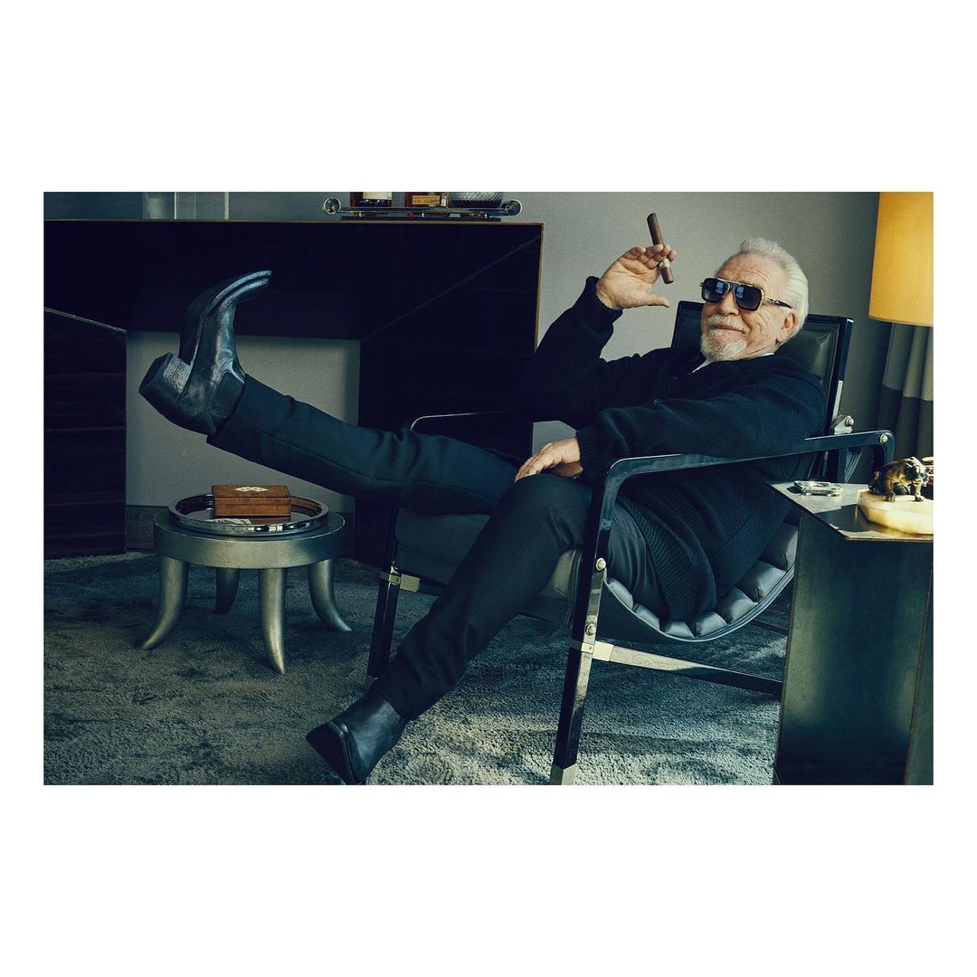 Brian Cox is relaxing in a chair with his feet up, holding a cigar in a photo studio setting, wearing black clothing and boots.