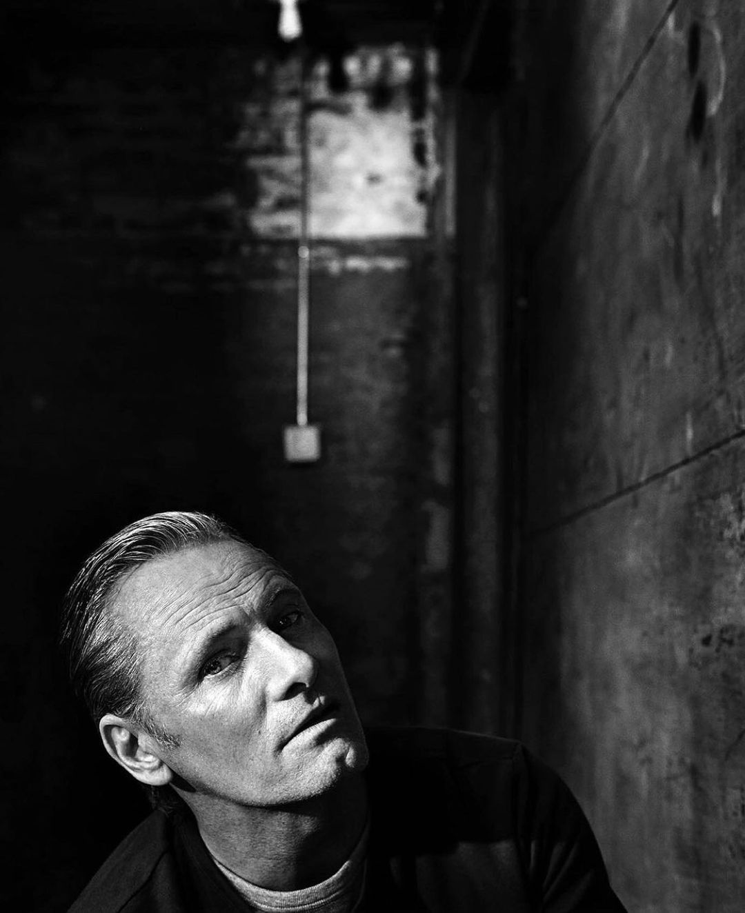 Black and white portrait of an adult male looking up in a dark indoor environment with industrial walls and overhead lighting.