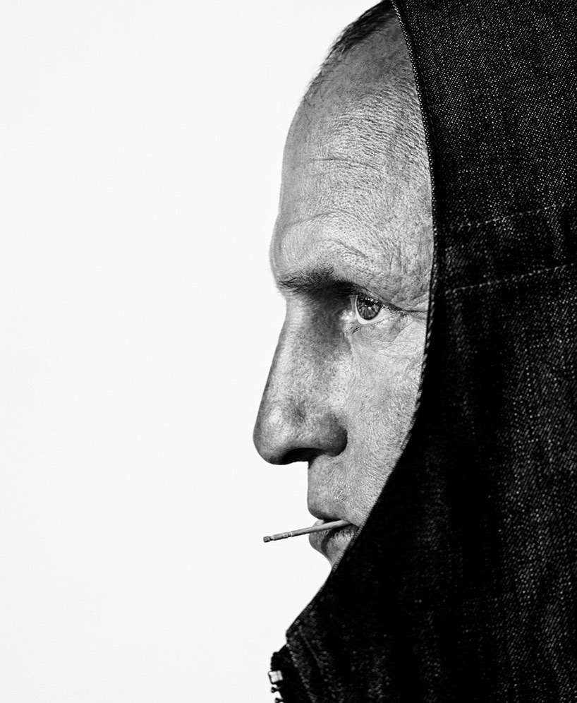 Portrait of an adult man in a dark hooded jacket with a matchstick in his mouth, set against a bright white background in black and white.