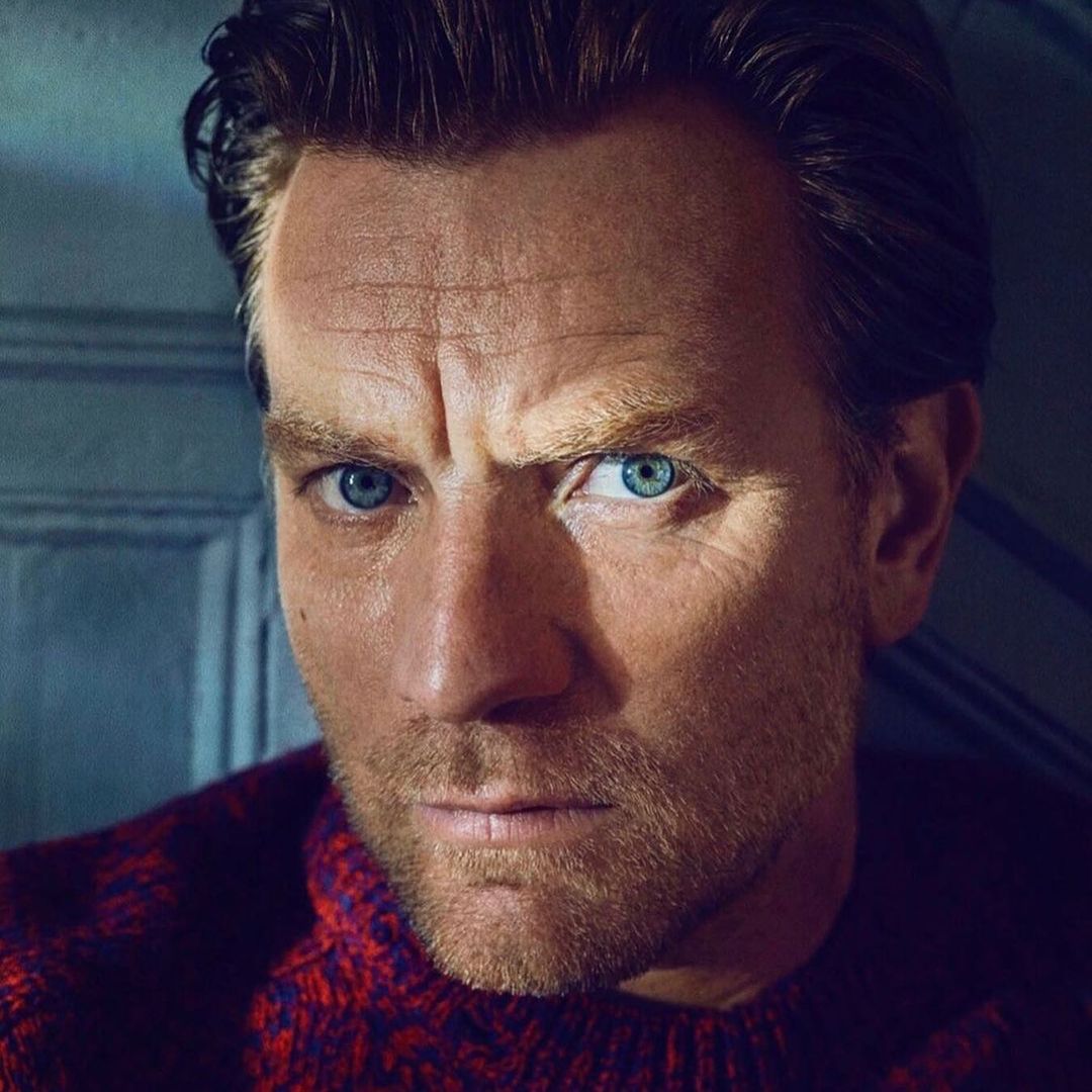 Close-up portrait of a fair-skinned adult male with blue eyes, wearing a patterned red and blue sweater, gazing intensely at the viewer.