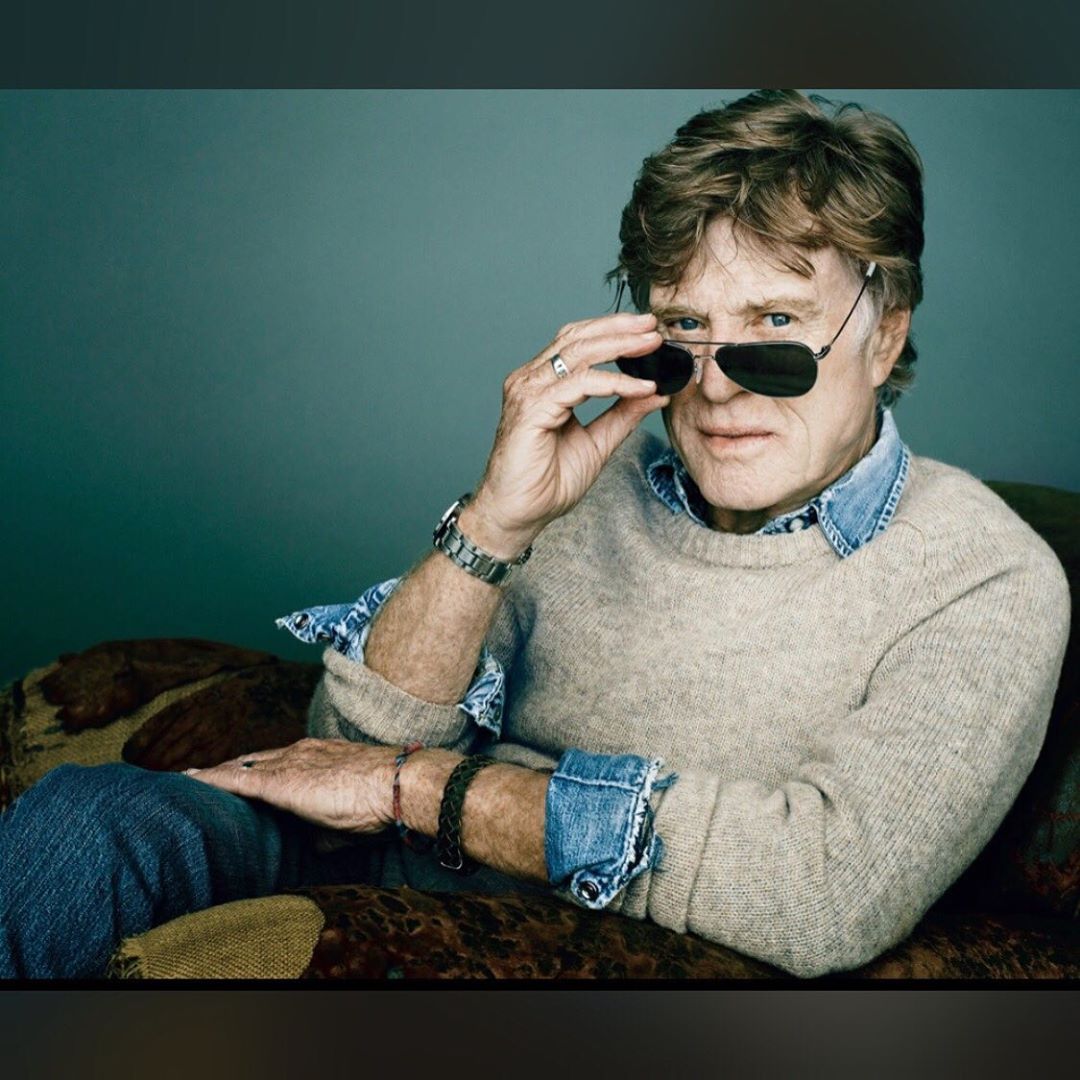 Portrait of an adult man, wearing sunglasses and a sweater, resting on a patterned couch against a solid-color background.