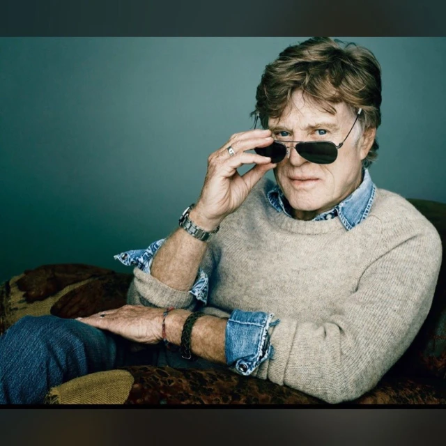 Portrait of an adult man, wearing sunglasses and a sweater, resting on a patterned couch against a solid-color background.