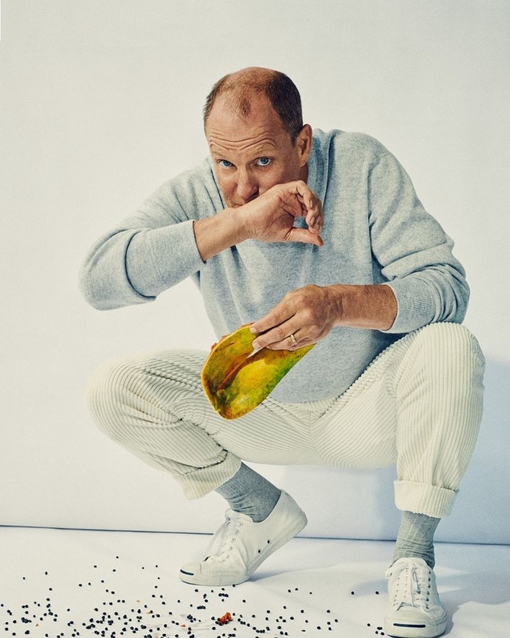 Portrait of adult man crouching and holding a taco in a studio setting, wearing a light grey sweatshirt, white pants and sneakers.