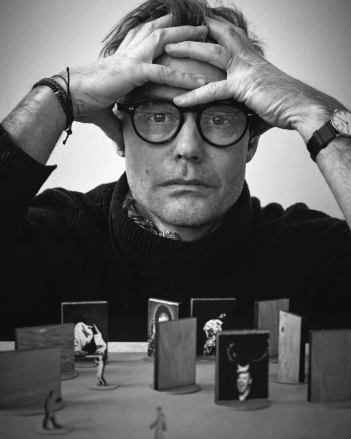 Monochrome portrait of a man with glasses holding his head surrounded by small figurine setups with photo prints, in a photo studio.