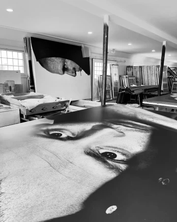 Monochrome photograph features a large-scale portrait and partial portrait in an artist's studio with windows and various studio equipment.