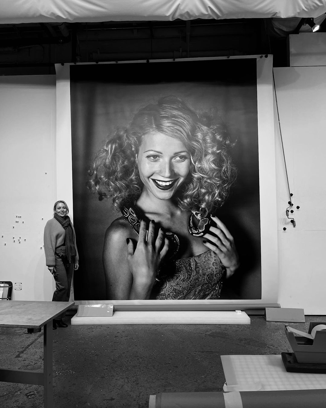 Black and white photo of a woman standing next to a large poster of a woman with a snake around her neck in a studio.