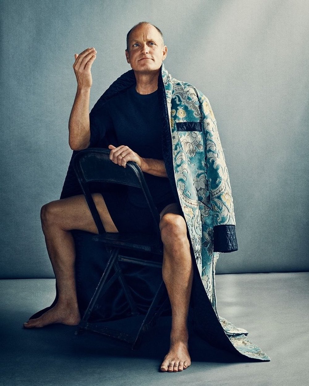 Portrait of Woody Harrelson sitting on a black folding chair in a studio, wearing a black shirt and a patterned turquoise robe.