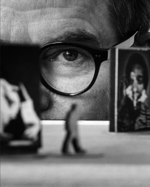 Close-up black and white portrait featuring a person's eye with glasses, and two blurred framed portraits in the background.