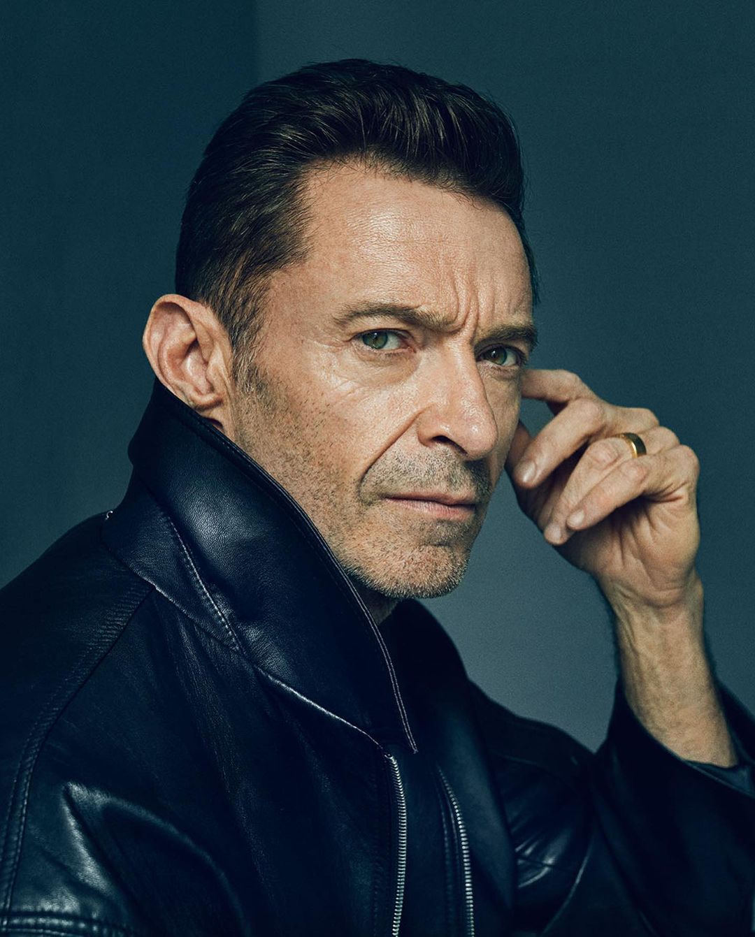 Close-up portrait of adult male with dark hair, wearing a black leather jacket, gazing at the camera against a gray background.